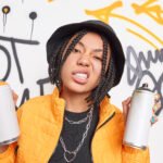 cheeky hipster girl with dreadlocks clenches teeth smirks face holds two aerosol sprays wears fashionable clothes draws graffiti wall has street lifestyle colorful manicure belongs to youth subculture