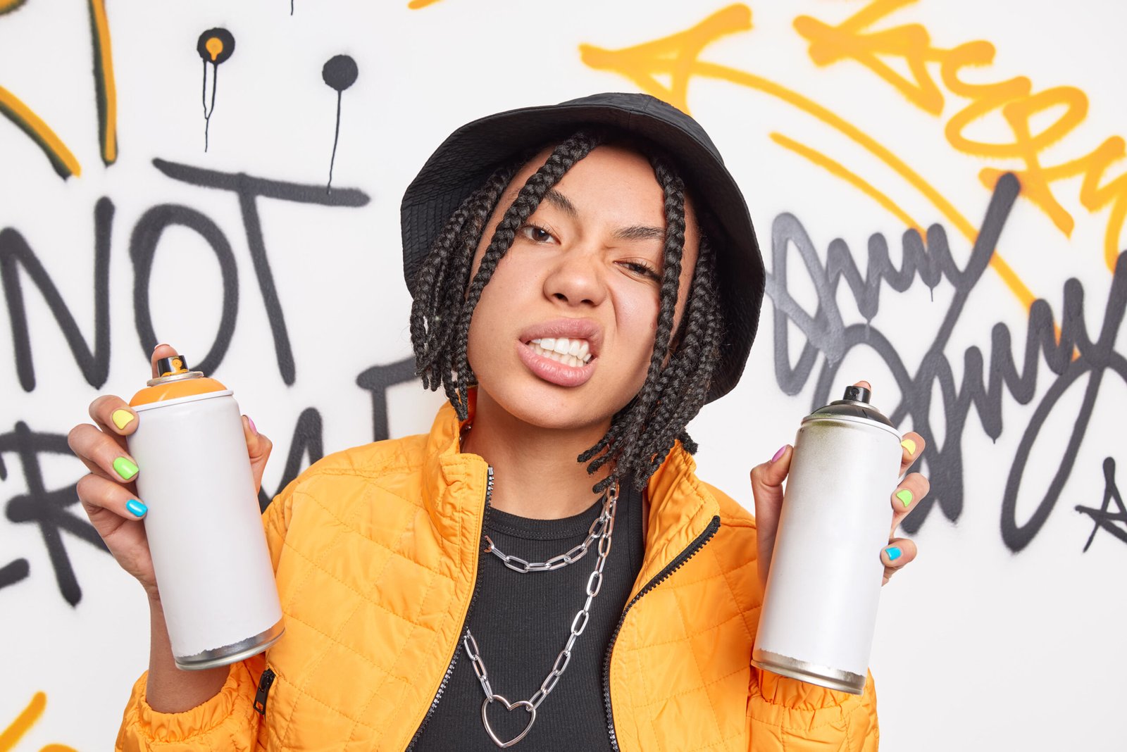 cheeky hipster girl with dreadlocks clenches teeth smirks face holds two aerosol sprays wears fashionable clothes draws graffiti wall has street lifestyle colorful manicure belongs to youth subculture