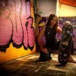Young woman kneeling with a skateboard in front of a colorful graffiti wall.