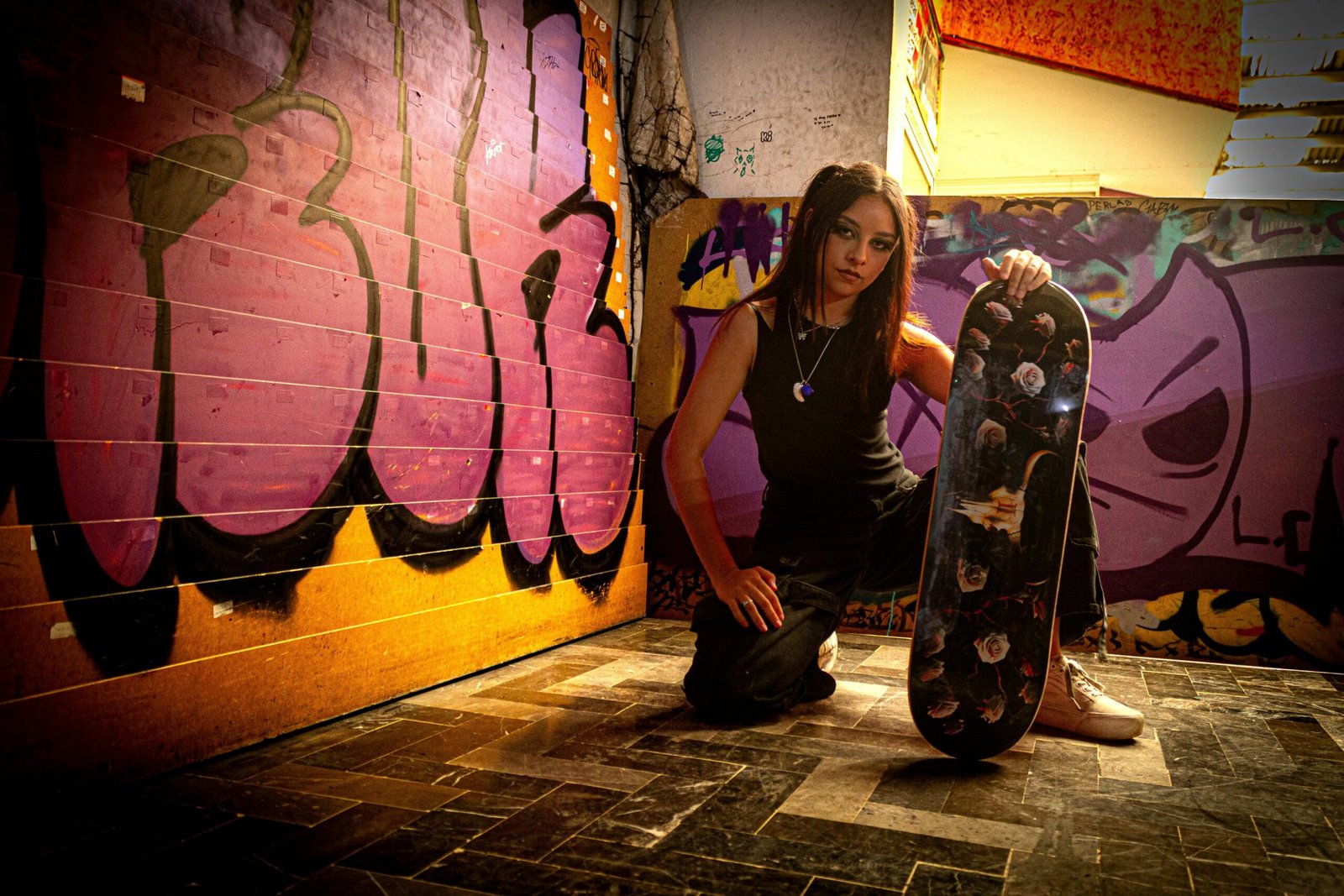 Young woman kneeling with a skateboard in front of a colorful graffiti wall.